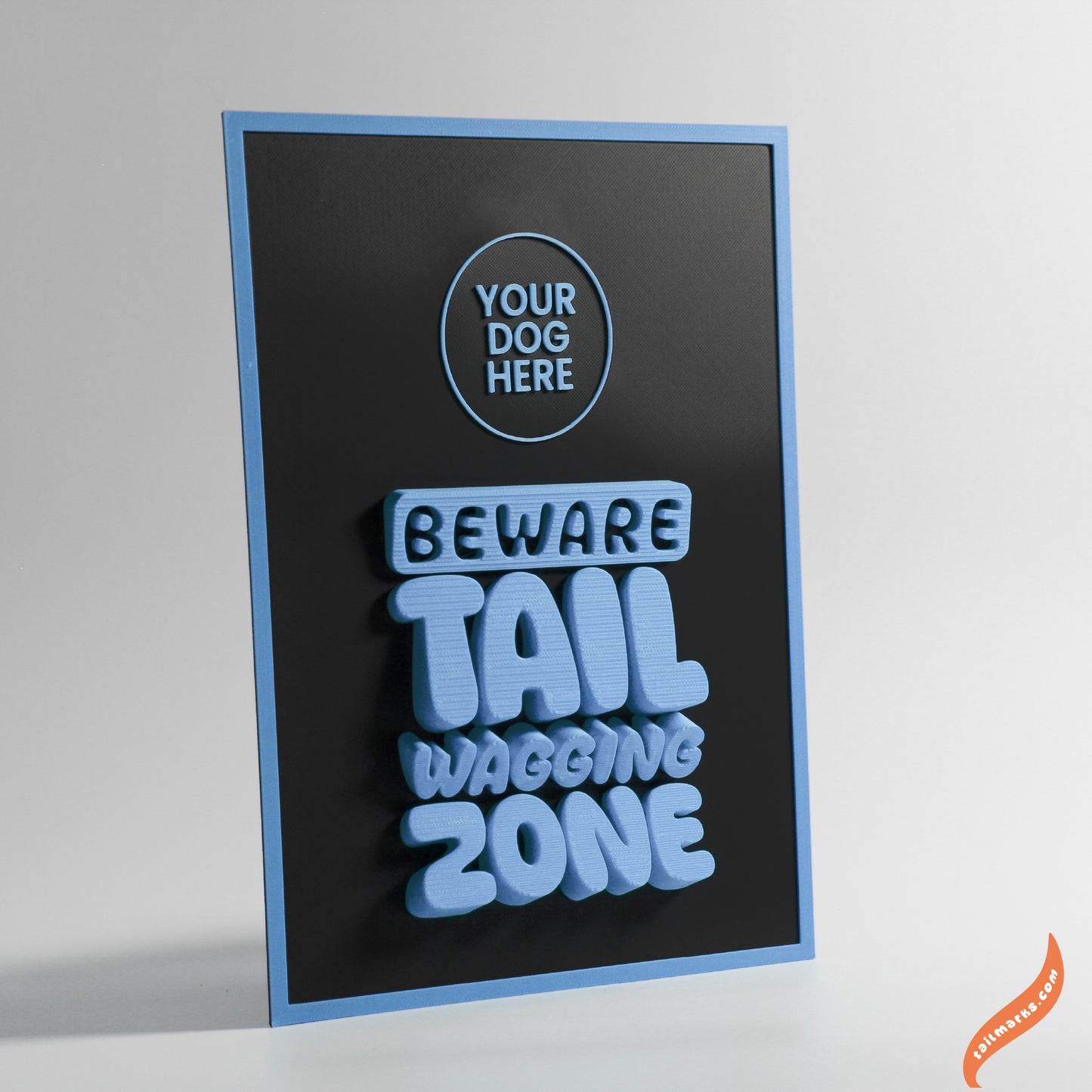 Black sign with blue text 'Beware Tail Wagging Zone' with a slim frame on a gray background