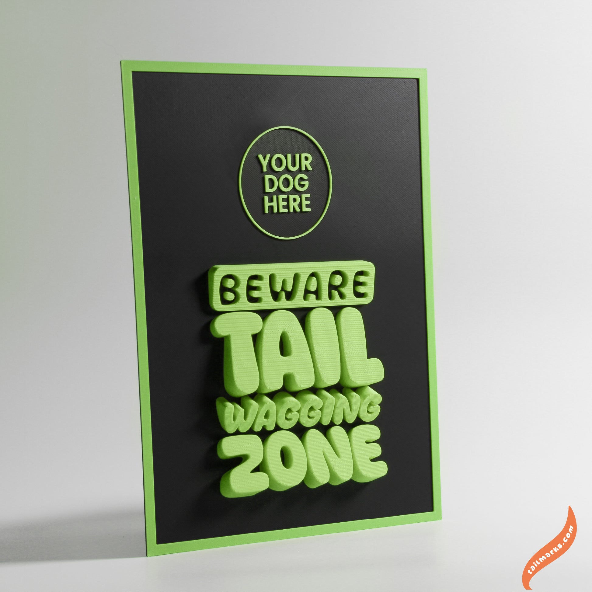 Black sign with green text 'Beware Tail Wagging Zone' and a slim frame on a white background